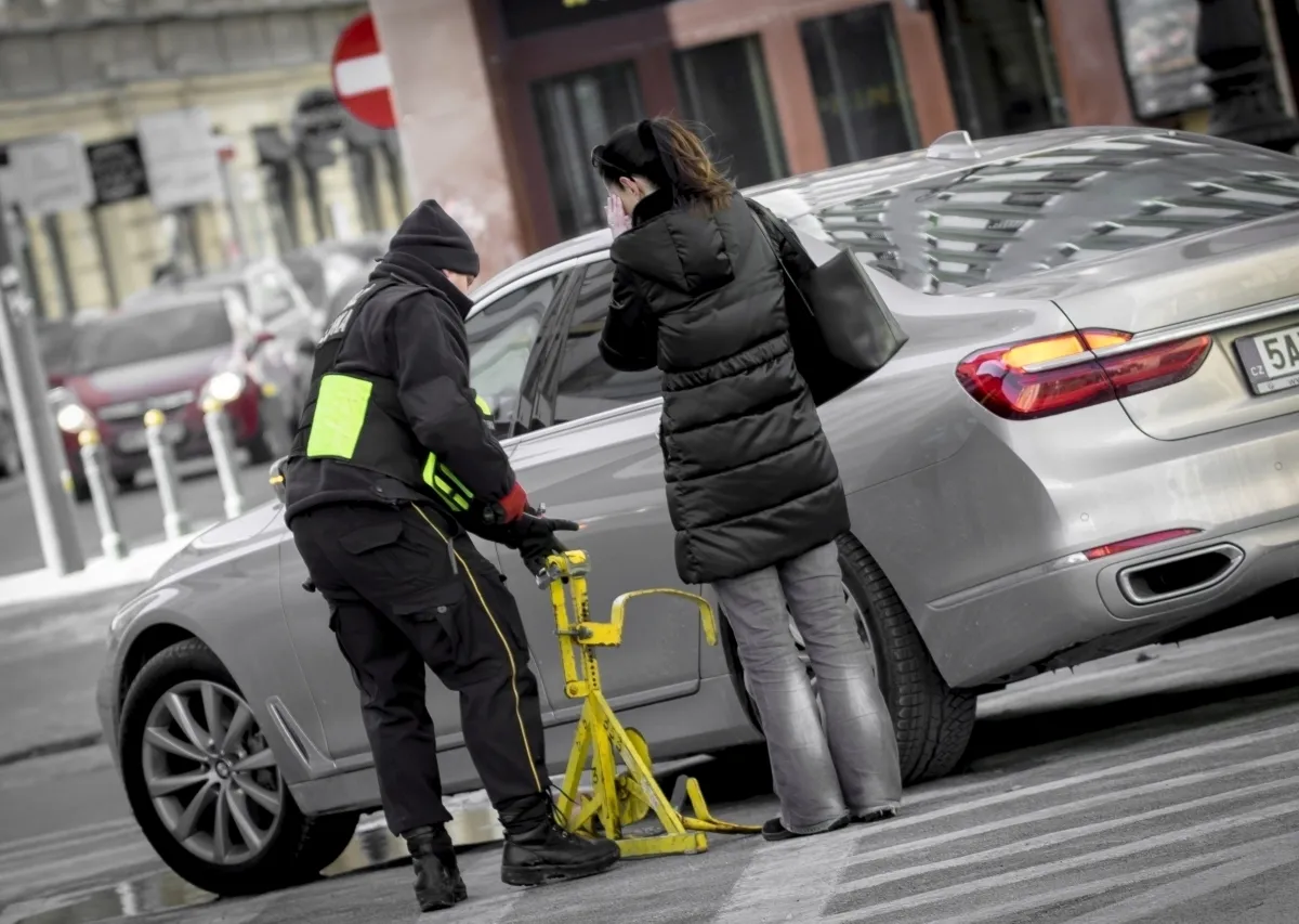 Parking officer positioning a wheel clamp beside a silver car while the driver speaks on a phone